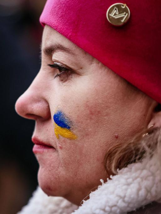 Profil einer Demonstrantin mit einer roten Mütze und einer gemalten ukrainischen Flagge auf der Wange.