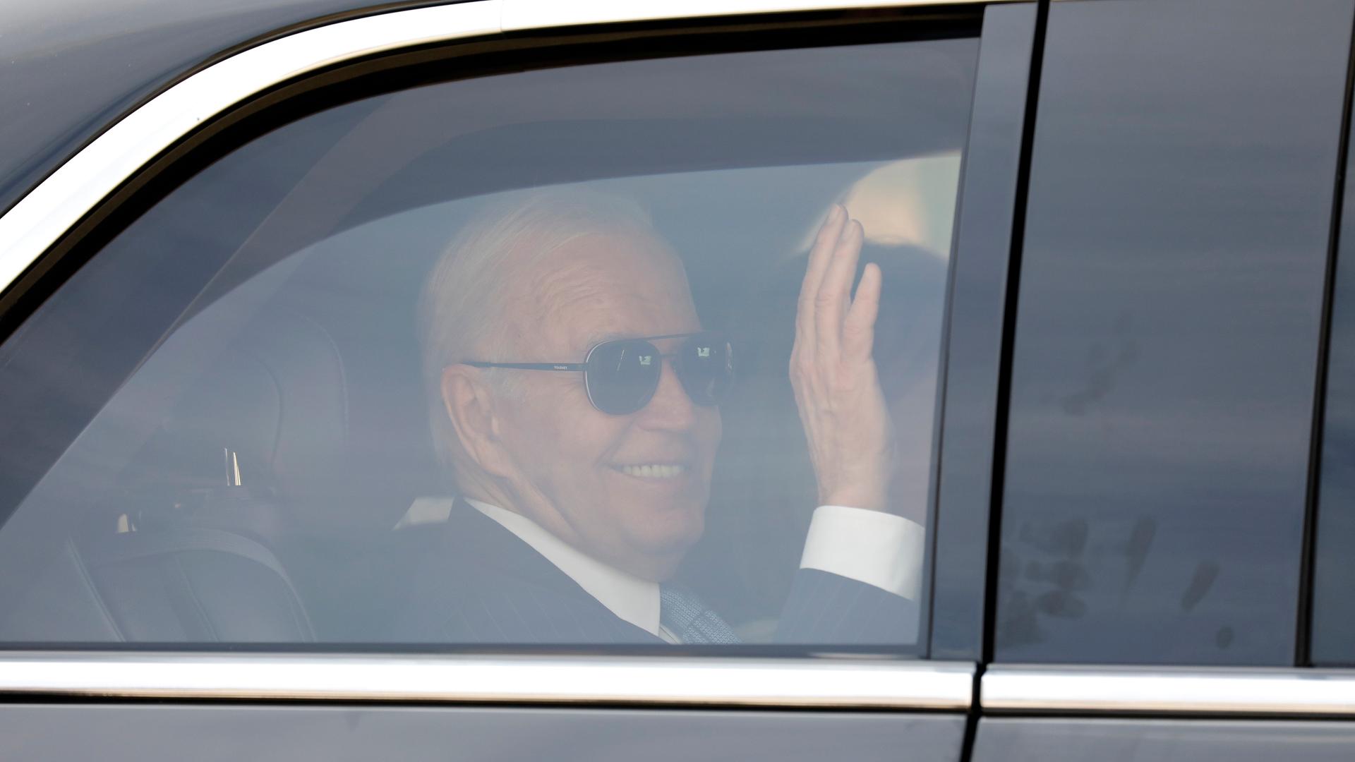 Joe Biden ist mit Sonnebrille hinter dem Fenster einer schwarzen Limousine zu sehen. Er lächelt und winkt.