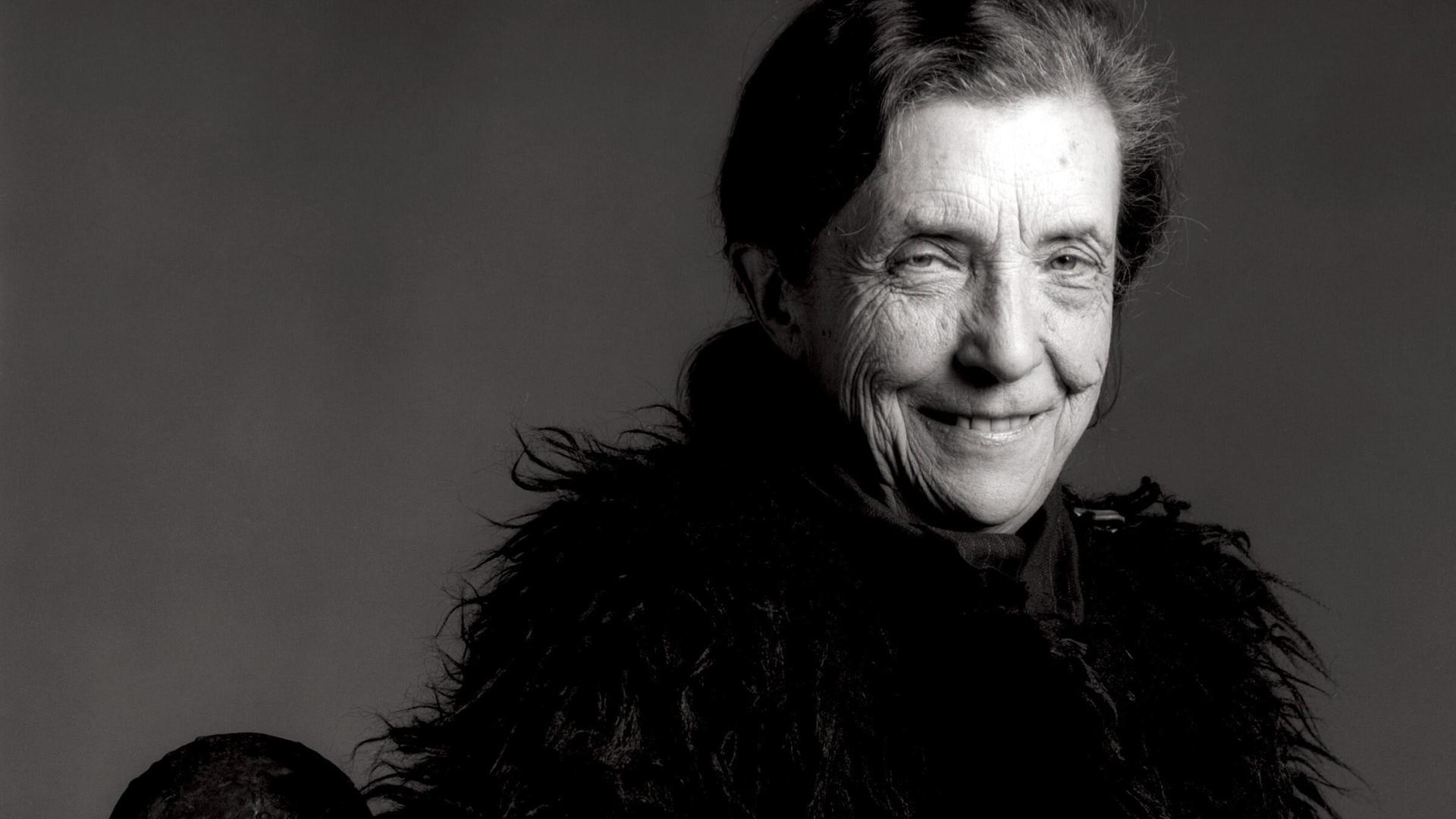 Ein Schwarz-Weiß-Foto von Louise Bourgeois, auf dem sie mit einer unter den Arm geklemmten überdimensionierten Penisskulptur zu sehen ist.