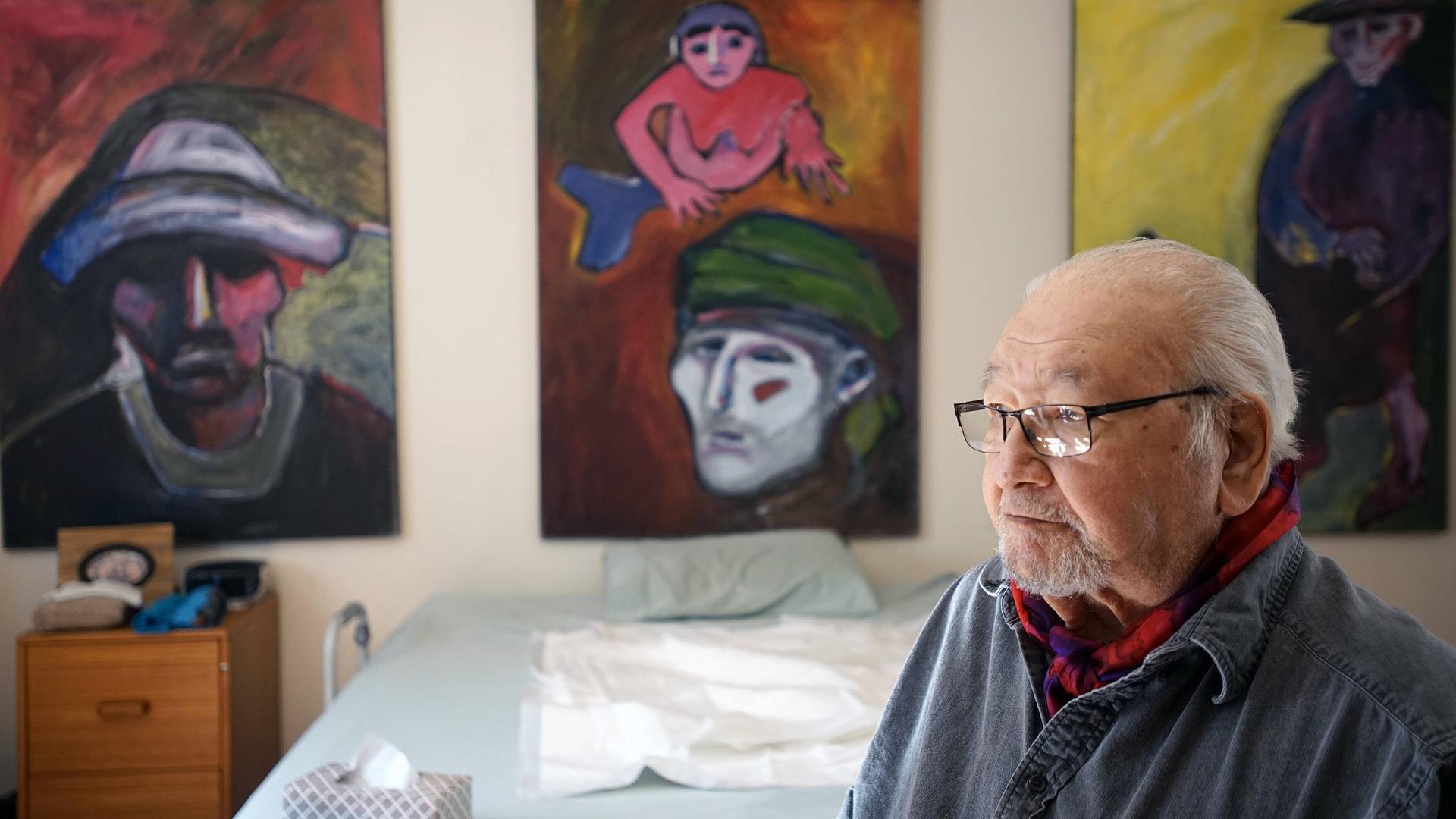 Ein älterer Mann mit Brille und grauen Haaren sitzt in einem Raum. Im Hintergrund an der Wand hängen Gemälde