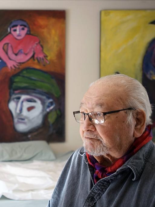 Ein älterer Mann mit Brille und grauen Haaren sitzt in einem Raum. Im Hintergrund an der Wand hängen Gemälde