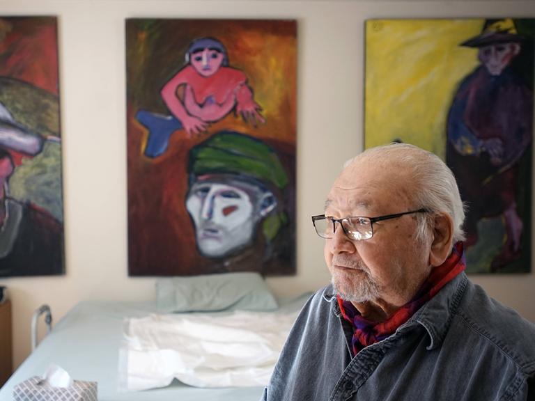 Ein älterer Mann mit Brille und grauen Haaren sitzt in einem Raum. Im Hintergrund an der Wand hängen Gemälde