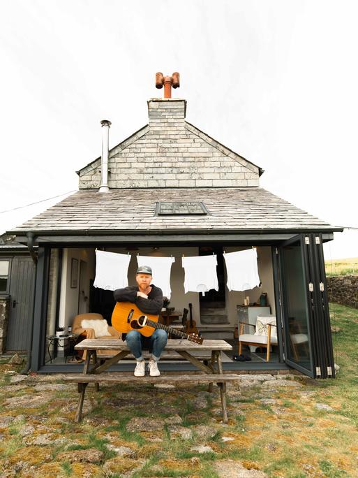 Stu Larsen sitzt vor einem kleinen, einsamen Haus auf einem Holztisch mit seiner Gitarre und schaut in die Kamera. 