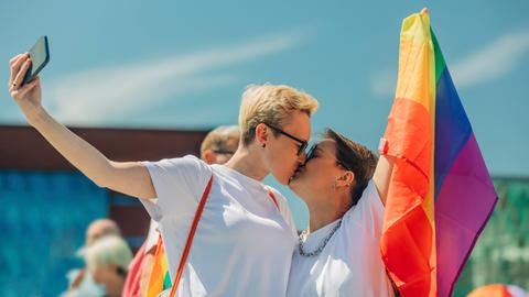 Zwei Frauen küssen sich, während sie eine Regenbogenfahne halten und ein Selfi machen.