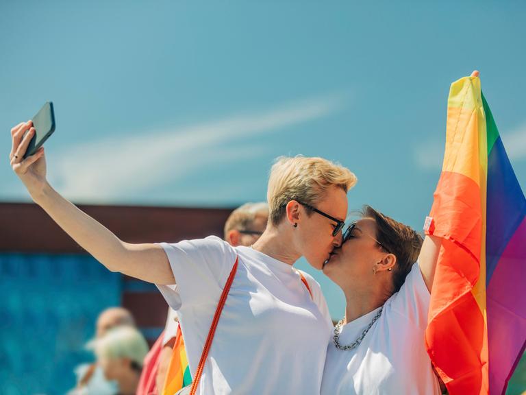 Zwei Frauen küssen sich, während sie eine Regenbogenfahne halten und ein Selfi machen.