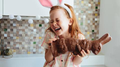 Ein rothaariges Mädchen hält einen Schokoosterhasen in der Hand und ist darüber sichtlich erfreut. Sie trägt einen Haarreifen mit Stoffohren und sieht damit selbst aus wie ein Osterhase.