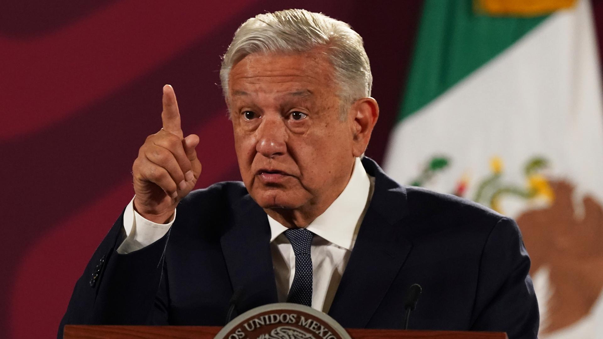Der Präsident spricht mit erhobenem Zeigefinger an einem Podest vor einer Landesflagge. Der Präsident spricht mit erhobenem Zeigefinger an einem Podest vor einer Landesflagge.