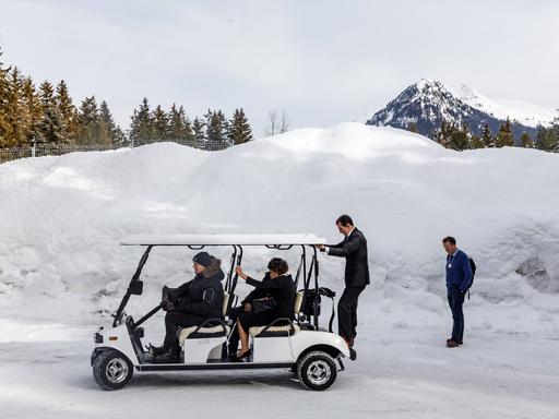 Teuer gekleidete Besucher des Weltwirtschaftsforums in Davos/Schweiz sitzen in einem Golf Trolley und werden durch den Schnee gefahren, im Hintergrund sind Berge zu sehen.
