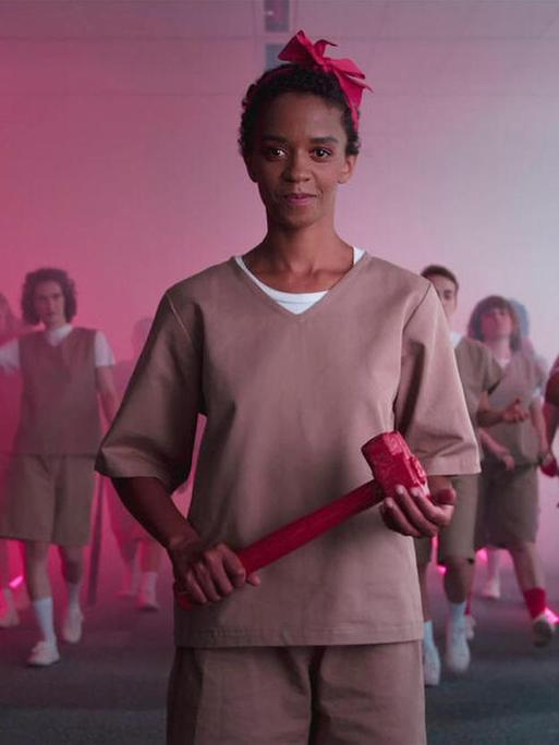 Im Still aus "Feminism WTF" schaut eine Schwarze Frau in altrosa Klamotten in die Kamera und hält einen pinken Hammer. Hinter ihr sind weitere Frauen mit pinken Utensilien wie Boxhandschuhen, Ketten und Werkzeugen versammelt.