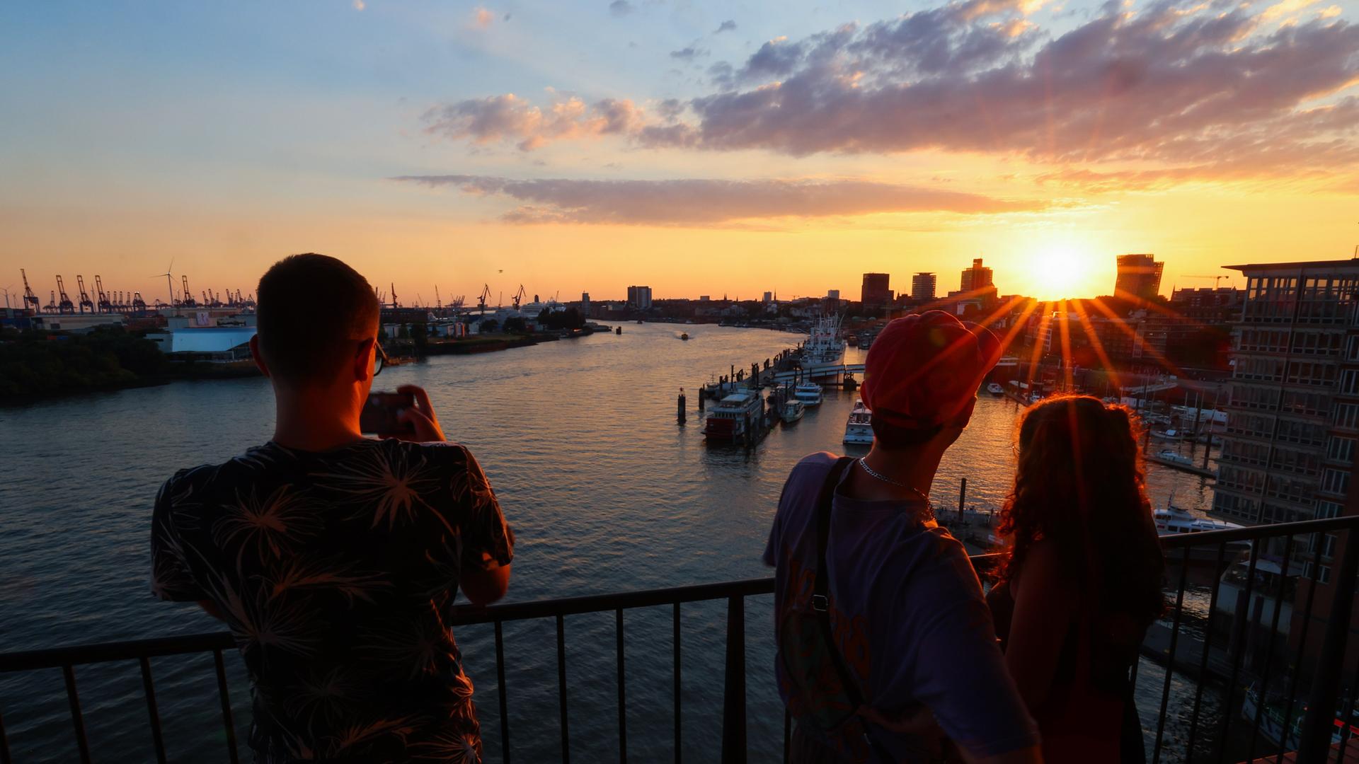 Menschen stehen auf der Plaza der Elbphilharmone und beobachten den Sonnenuntergang über der Elbe und den Landungsbrücken.
