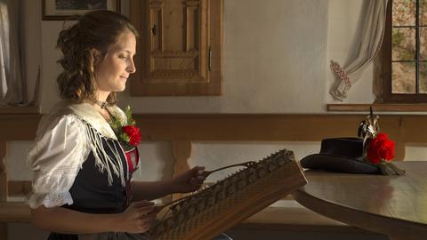 Eine junge Frau sitzt in Tracht in einer bäuerlichen Stube und spielt auf einem Hackbrett.