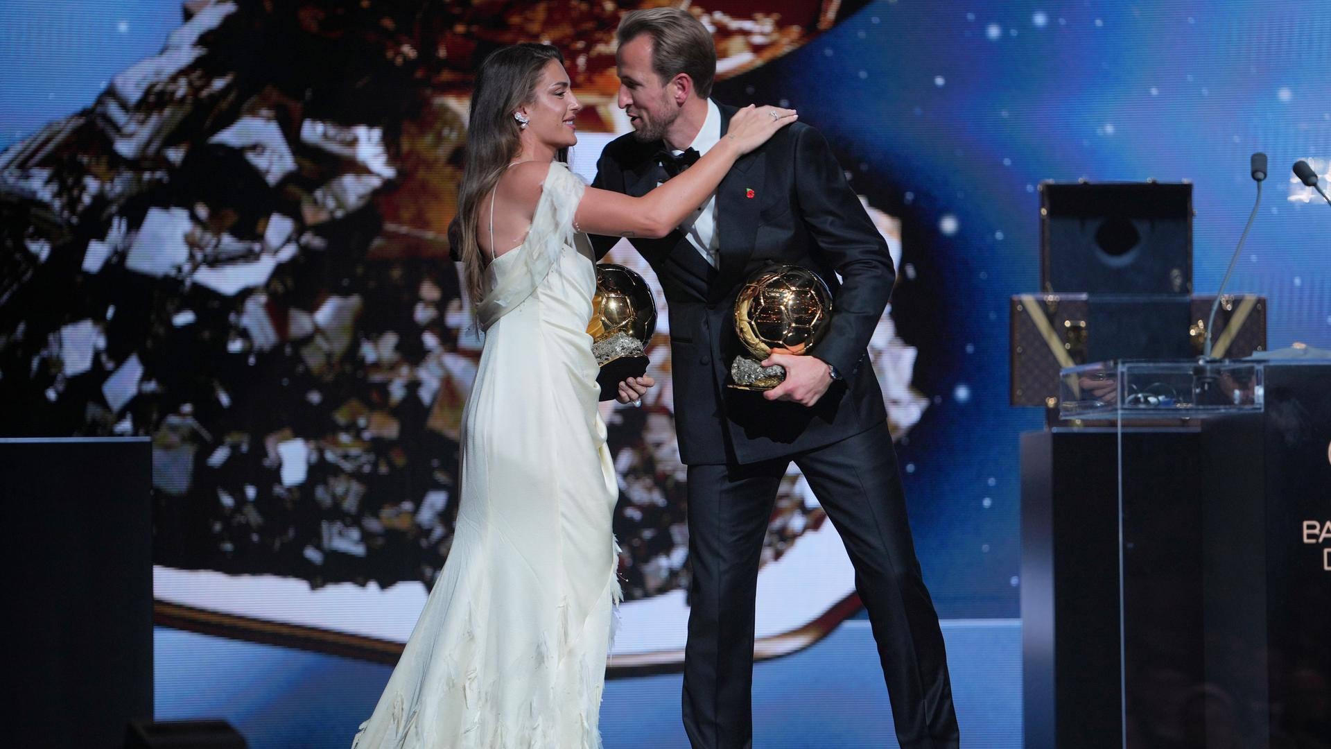 Harry Kane und Alexia Putellas stehen in Abenkleidung auf der Bühne und beugen sich zum Küsschen einander zu. Beide halten jeweils den Ballon d'Or in der Hand. Harry Kane und Alexia Putellas stehen in Abenkleidung auf der Bühne und beugen sich zum Küsschen einander zu. Beide halten jeweils den Ballon d'Or in der Hand.