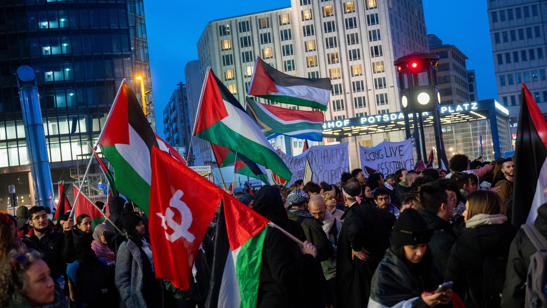 Teilnehmer einer Pro-Palästina-Demonstration unter dem Namen "Global South Resists" stehen auf dem Potsdamer Platz und halten Palästina-Fahnen in die Luft. Es dämmert. 