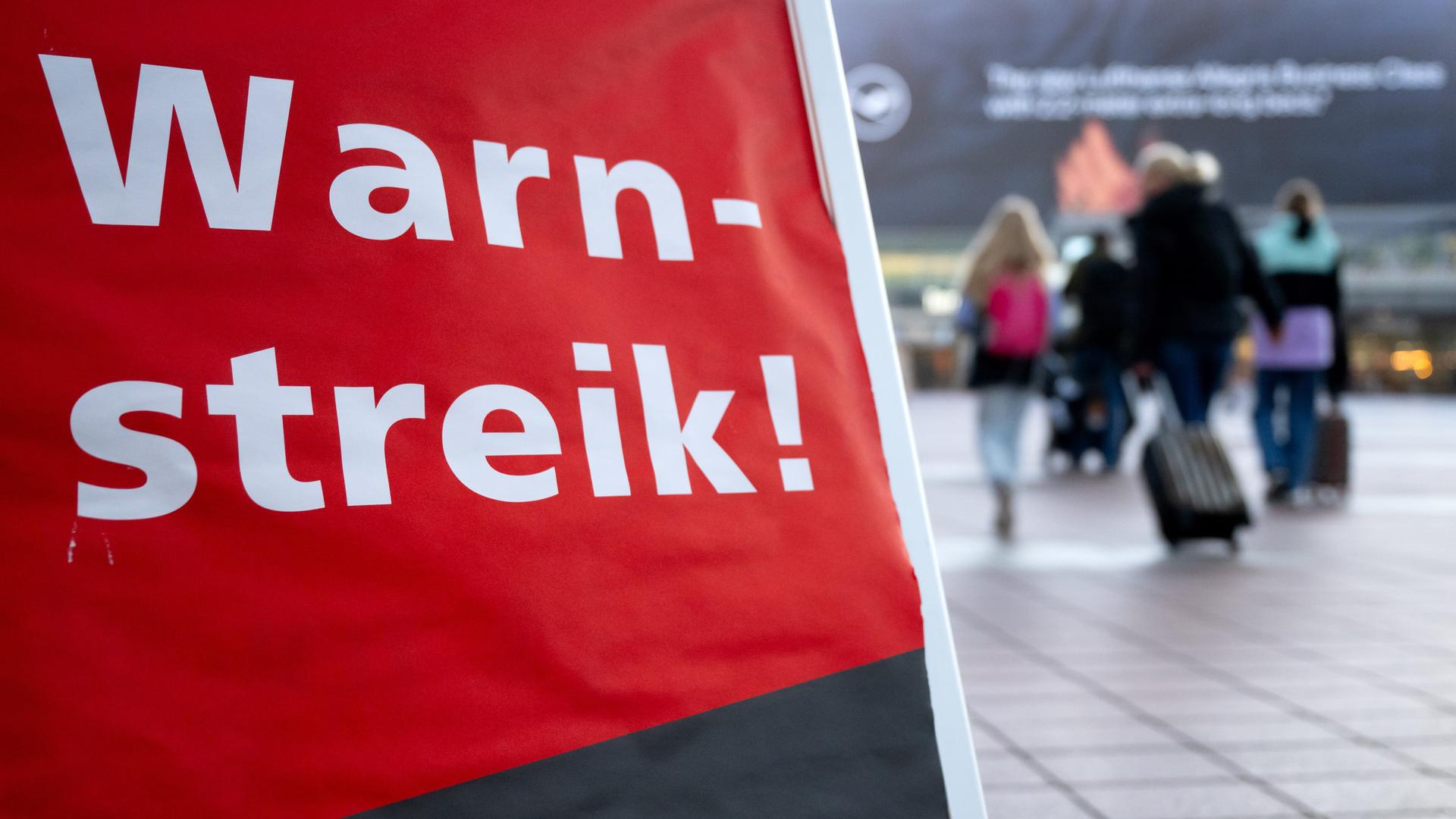 Passagiere gehen am Flughafen München mit ihrem Gepäck an einem Plakat mit der Aufschrift "Warnstreik" entlang zum Terminal. Passagiere gehen am Flughafen München mit ihrem Gepäck an einem Plakat mit der Aufschrift "Warnstreik" entlang zum Terminal.