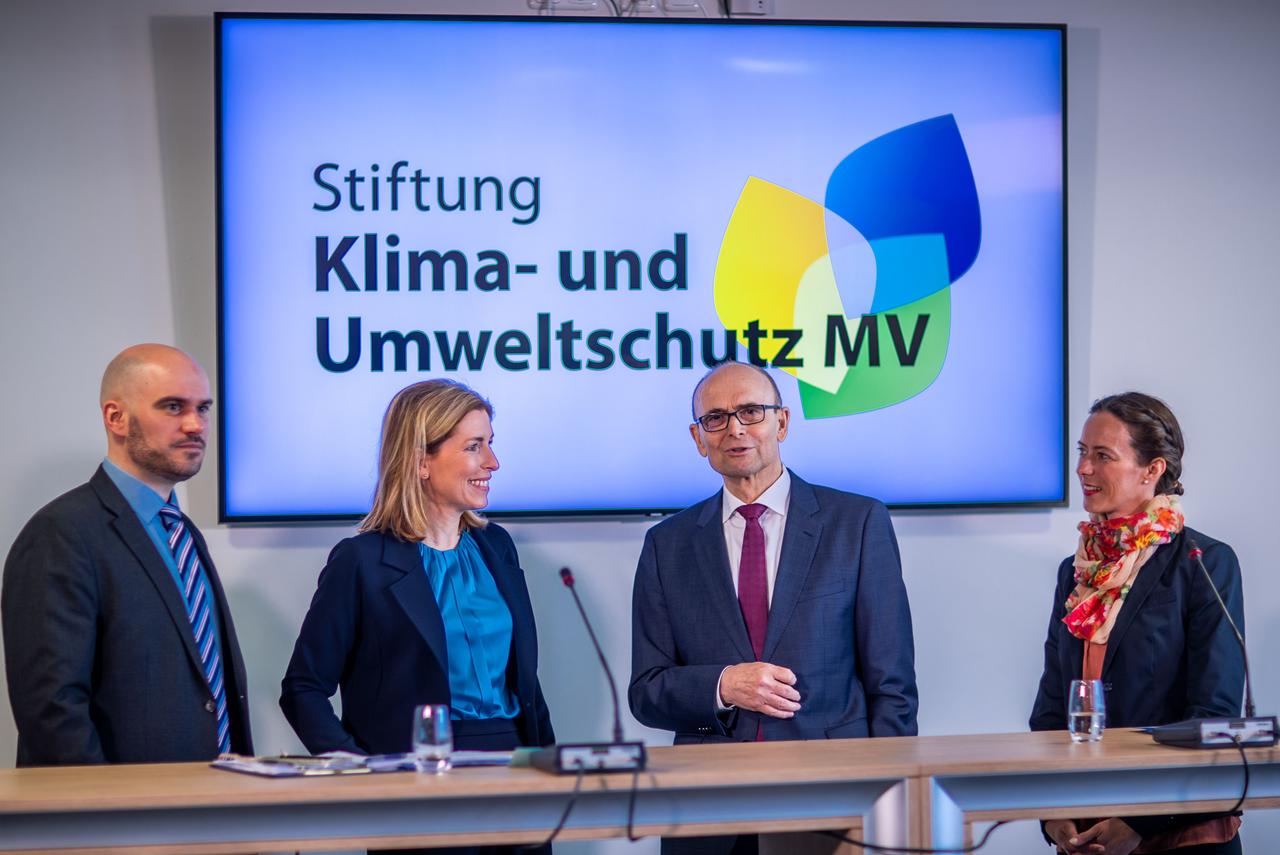 Vertreter der Stiftung Klima- und Umweltschutz Mecklenburg-Vorpommern bei einem Pressetermin. Ganz rechts die Geschäftsführerin, links von ihr Erwin Sellering. Im Hintergrund hängt ist auf einem Großbildschirm der Name der Stiftung und ihr Logo auf hellblauem Grund zu sehen. 