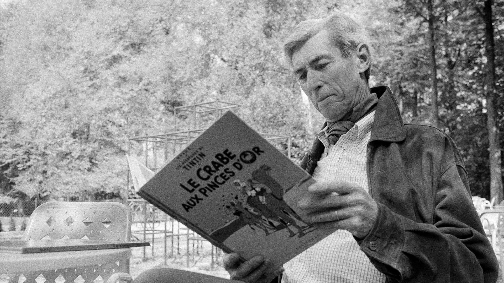 Hergé sitzt mit einer "Tim und Struppi"-Ausgabe in der Hand in einem Garten.