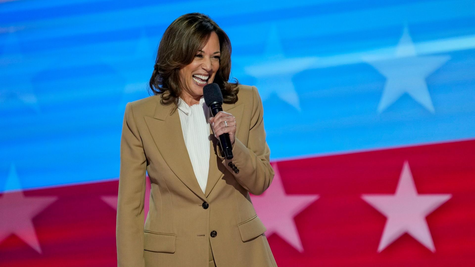 Kamala Harris mit einem Mikrophon in der Hand, hinter ihr auf einer Leinwand ein Ausschnitt der amerikanischen Flagge in blau und rot mit Sternen