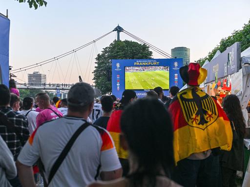 Menschen stehen mit Deutschland-Kleidung vor einem großen Bildschirm auf der Fanzone in Frankfurt.