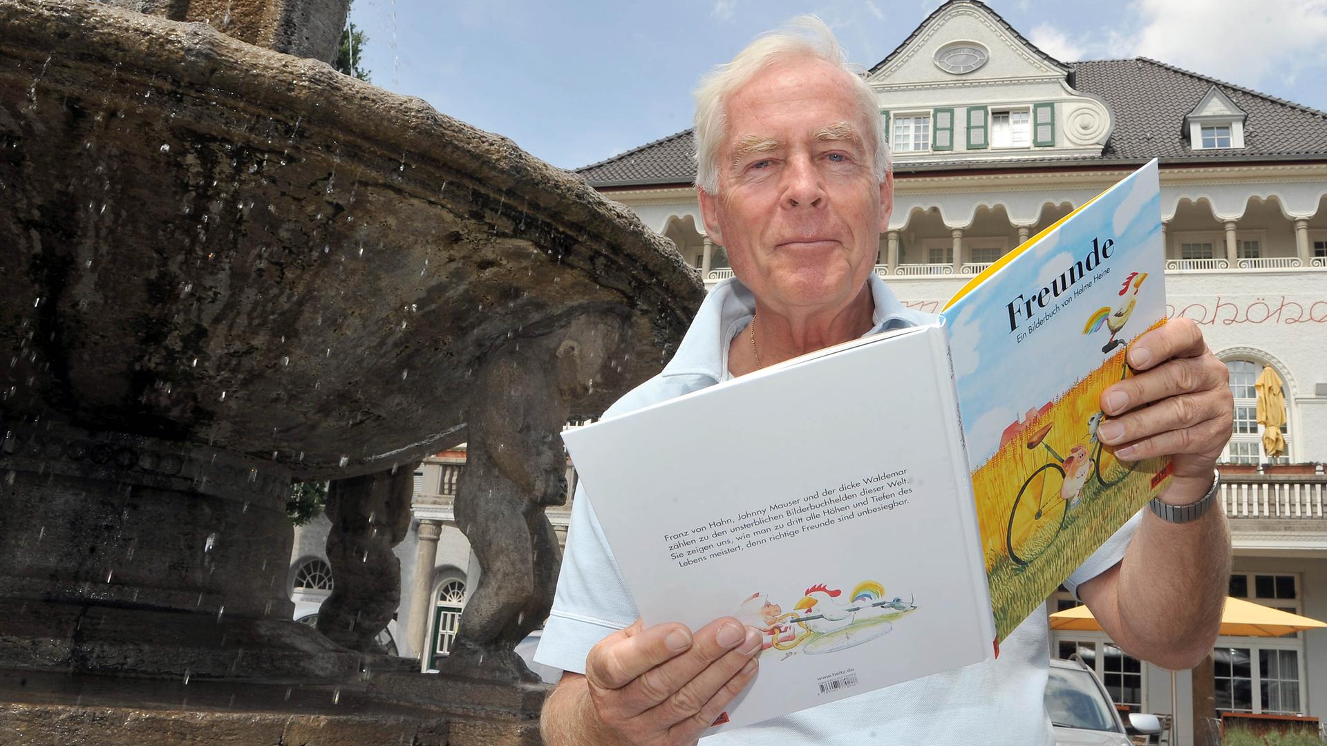 Der Kinderbuchautor und Illustrator Helme Heine vor einem Springbrunnen in Essen mit einer Ausgabe seines Kinderbuchklassikers "Freunde" aufgeschlagen in der Hand.