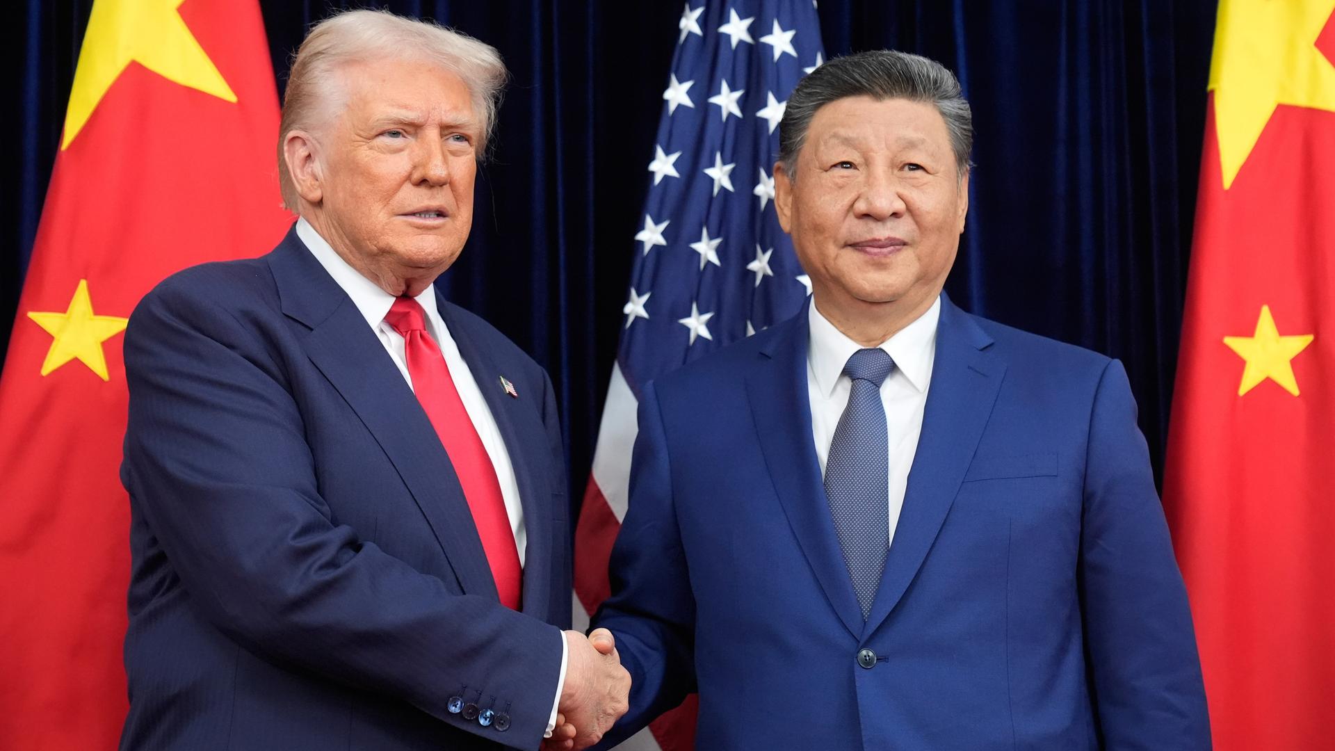 Donald Trump und Xi Jinping schütteln sich vor Flaggen beider Länder die Hände. Beide schauen dabei ernst.