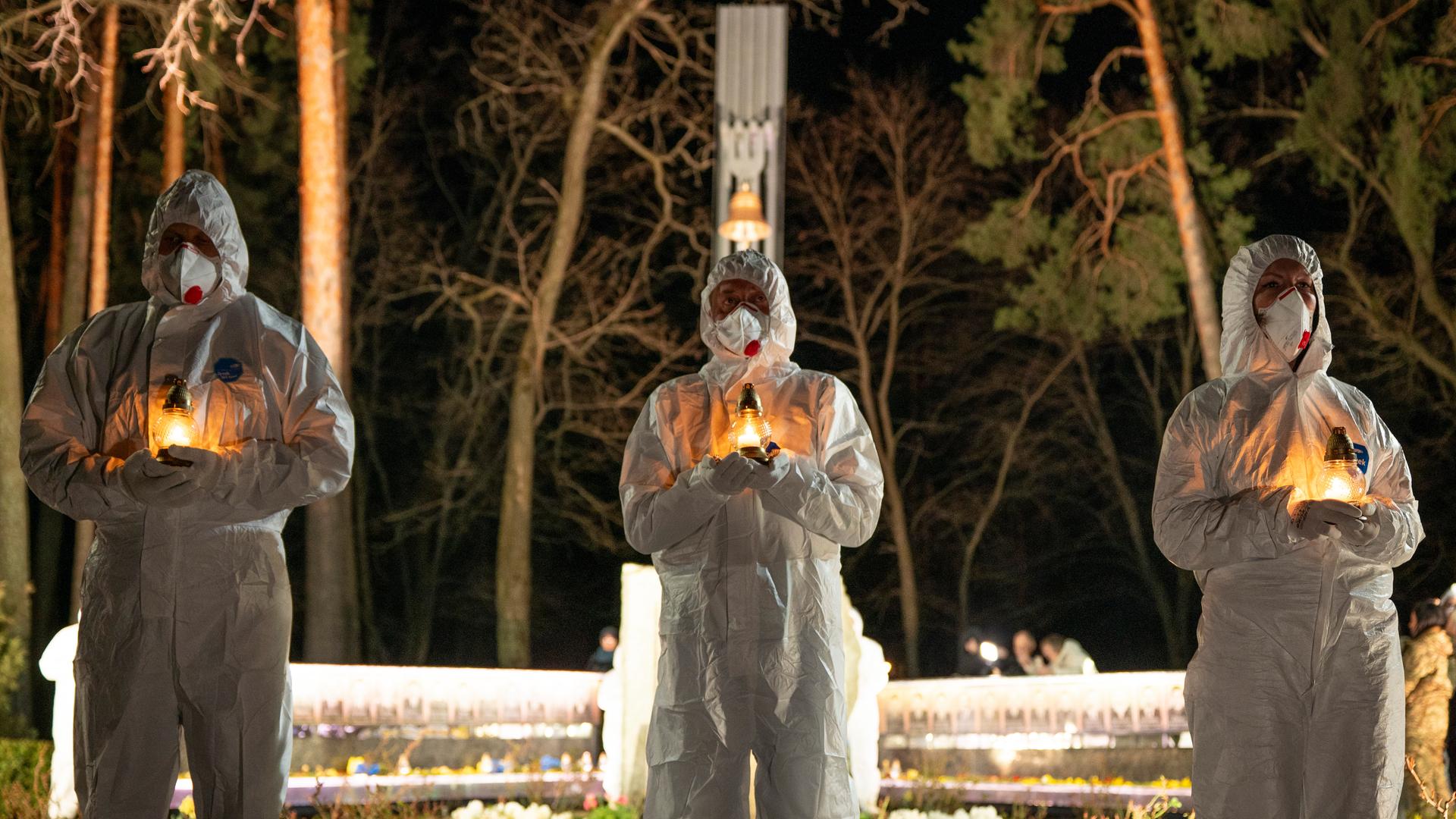 Menschen in weißen Schutzanzügen halten Kerzen vor einer Gedenkstätte. 