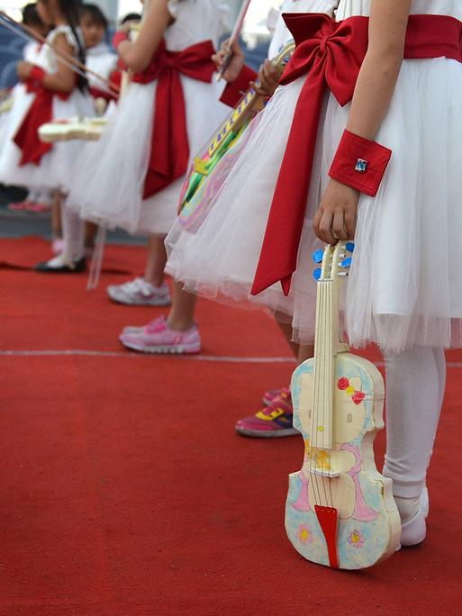 Junge Kinder in weiß-roten Kleidern stehen in Reihe und warten auf ihren Auftritt, denn ihre bunt bemalten Instrumente halten alle locker und noch nicht in Spielhaltung.
