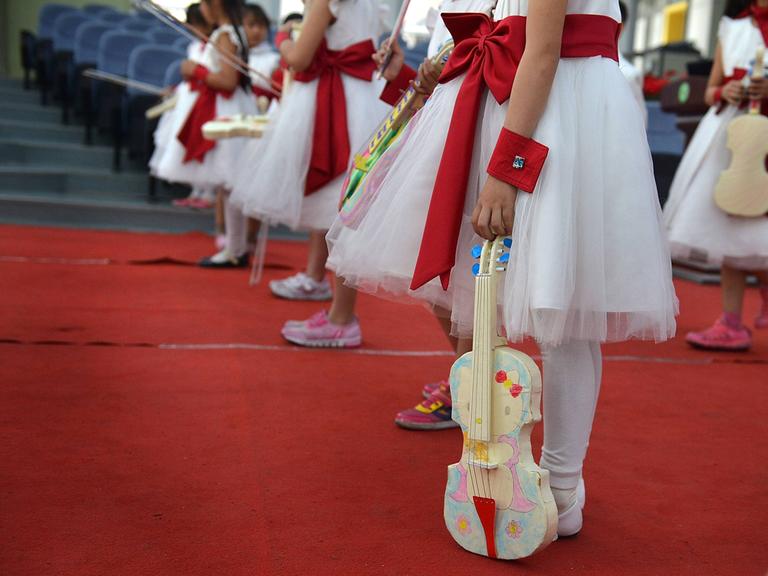 Junge Kinder in weiß-roten Kleidern stehen in Reihe und warten auf ihren Auftritt, denn ihre bunt bemalten Instrumente halten alle locker und noch nicht in Spielhaltung.