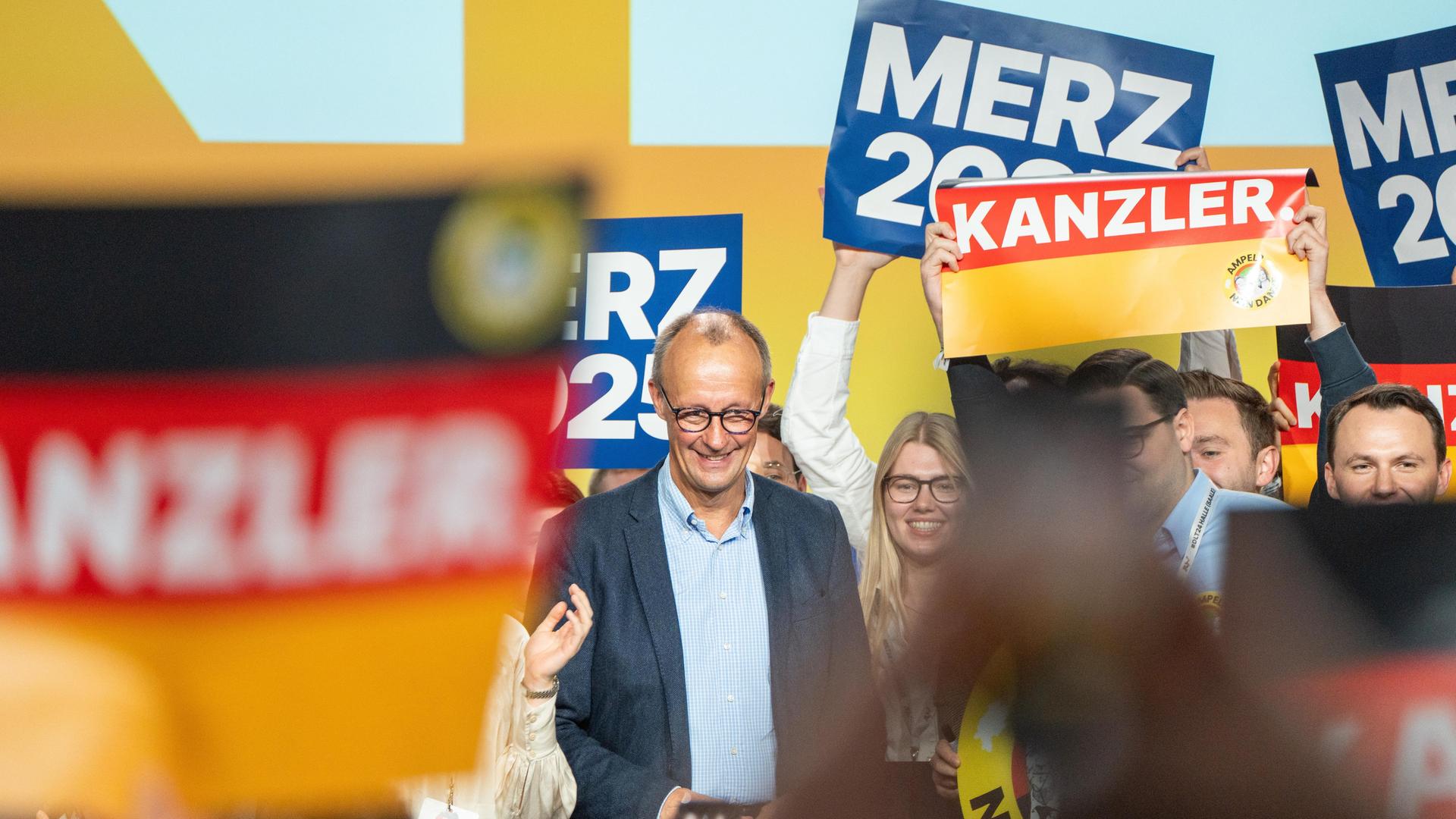 Friedrich Merz steht lächelnd auf einer Bühne, um ihn herum sieht man Plakate mit der Aufschrift "Merz 2025" und "Kanzler"