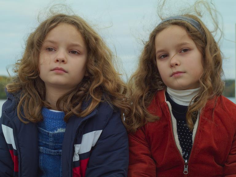 Zwei achtjährige Schwestern mit braunen Locken sitzen nebeneinander und blicken in die Kamera.