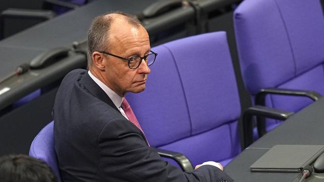 Friedrich Merz sitzt mit seitlich gedrehtem Oberkörper auf einem Stuhl im Plenarsaal des Bundestags. Sein Gesichtsausdruck wirkt skeptisch und müde. 