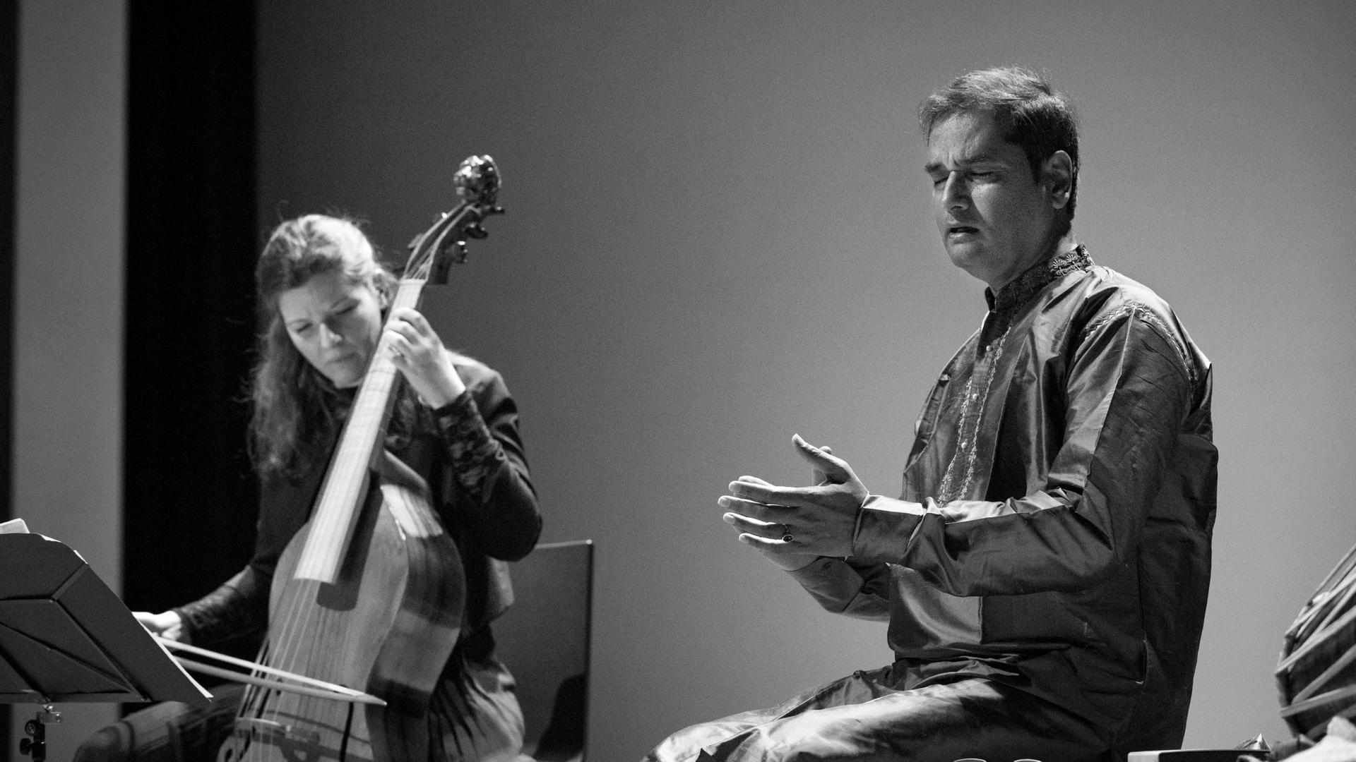 Eine Frau spielt auf einem Streichinstrument, einer Gambe, rechts neben ihr sitzt ein Mann und singt. Beide wirken versunken in die Musik. Das Foto ist in Grautönen aufgenommen.