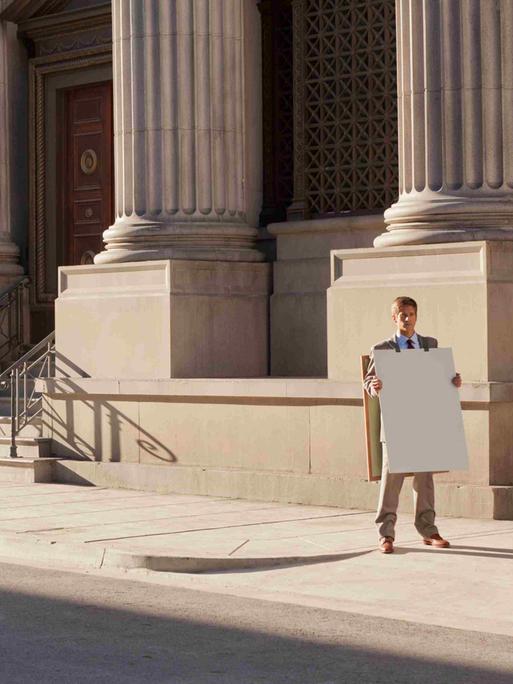 Ein Mann steht mit einem unbeschriebenen Sandwich-Board vor einem repräsentativen Gebäude mit Säulen.