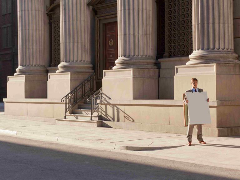 Ein Mann steht mit einem unbeschriebenen Sandwich-Board vor einem repräsentativen Gebäude mit Säulen.