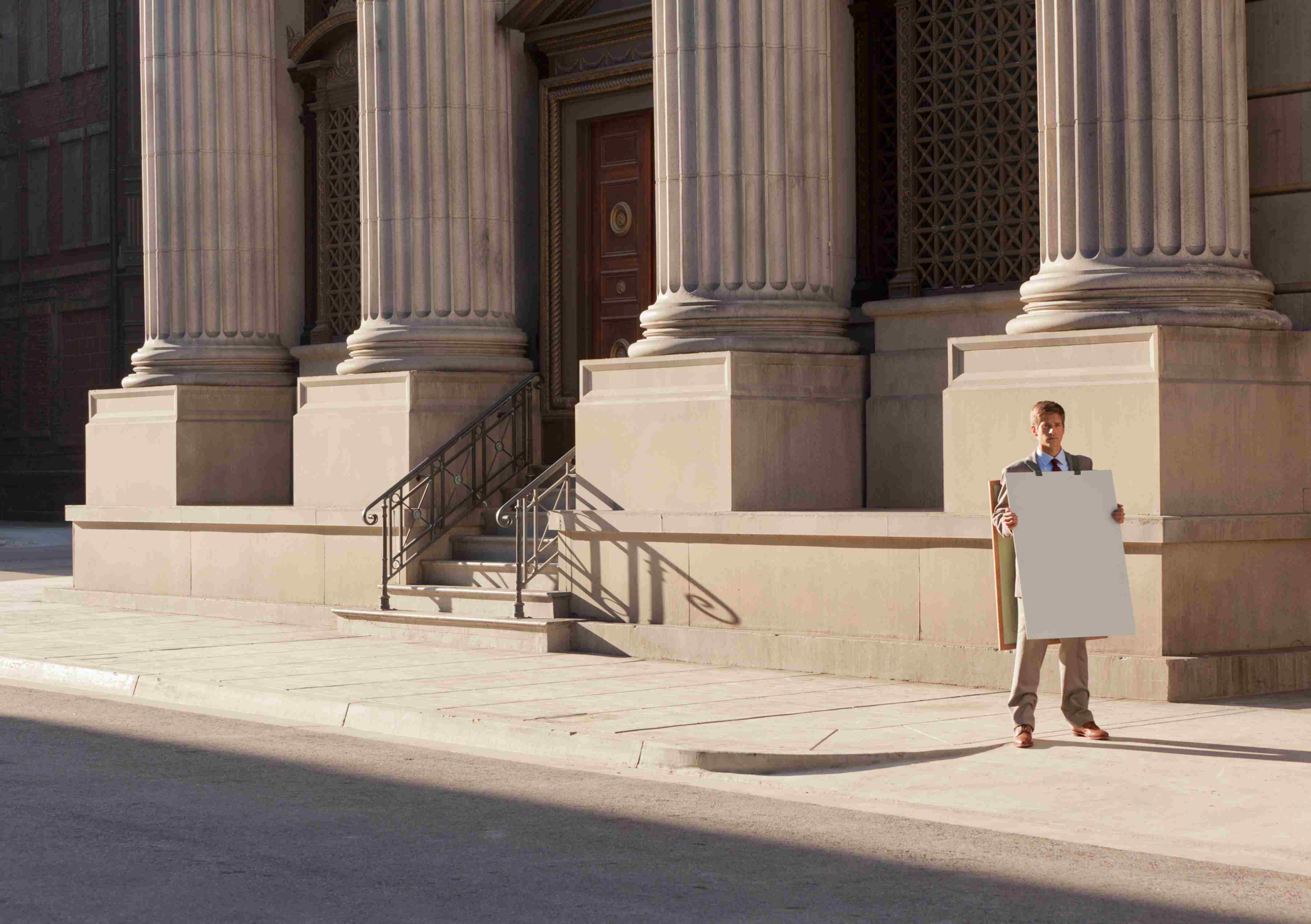 Ein Mann steht mit einem unbeschriebenen Sandwich-Board vor einem repräsentativen Gebäude mit Säulen.
