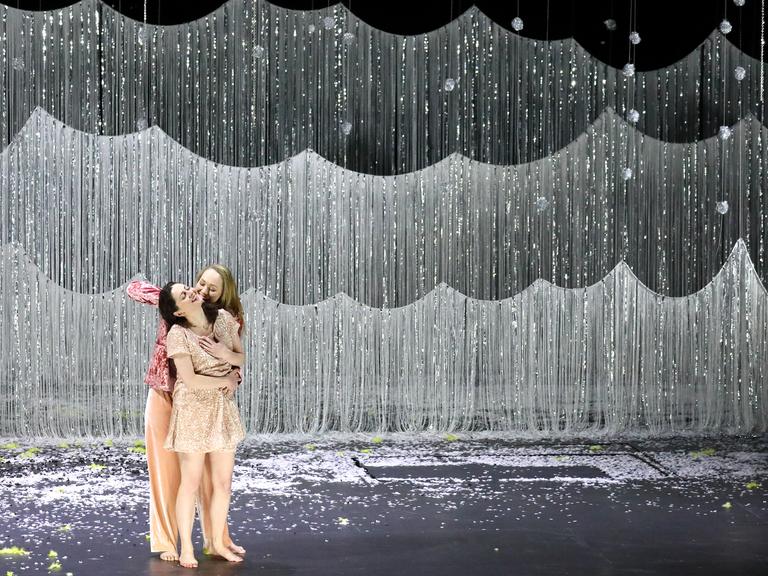 Zwei Frauen umarmen sich auf einer Bühne. Im Hintergrund hängt ein Glitzervorhang als Wald.