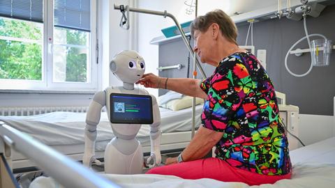 Eine ältere Dame sitzt auf einem Pflegebett und streicht einem humanoiden Roboter, der vor ihr steht, mit der rechten Hand über die Wange.