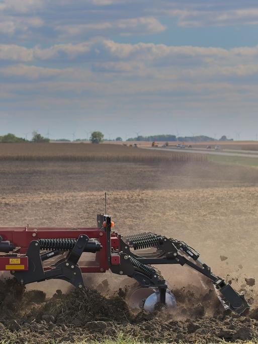 Eine rote Scheibenegge pflügt durch ein Feld und wirbelt Staub auf, während sie den Boden für die Aussaat vorbereitet. Die weitläufige Landschaft und die entfernte Baumgrenze umrahmen die Maschine unter einem teilweise bewölkten Himmel.
