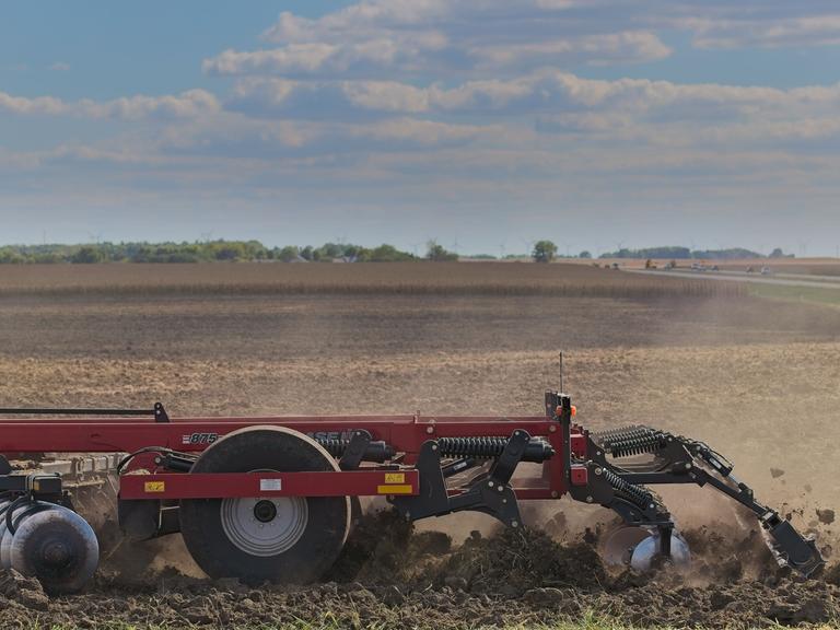 Eine rote Scheibenegge pflügt durch ein Feld und wirbelt Staub auf, während sie den Boden für die Aussaat vorbereitet. Die weitläufige Landschaft und die entfernte Baumgrenze umrahmen die Maschine unter einem teilweise bewölkten Himmel.