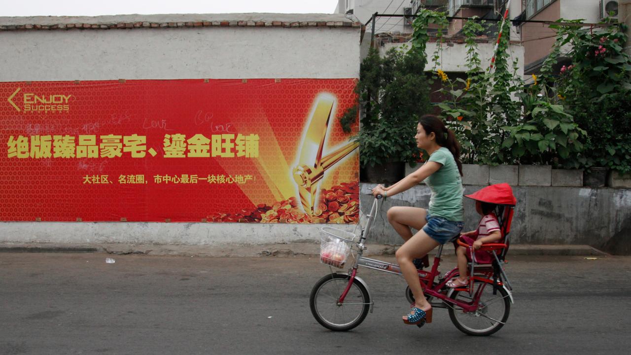 Eine Frau fährt in China auf einem kleinen Fahrrad eine Straße entlang, ein Kind sitzt hinten unter einem roten Sonnenschutz.
Eine Frau fährt in China auf einem kleinen Fahrrad eine Straße entlang, ein Kind sitzt hinten unter einem roten Sonnenschutz.