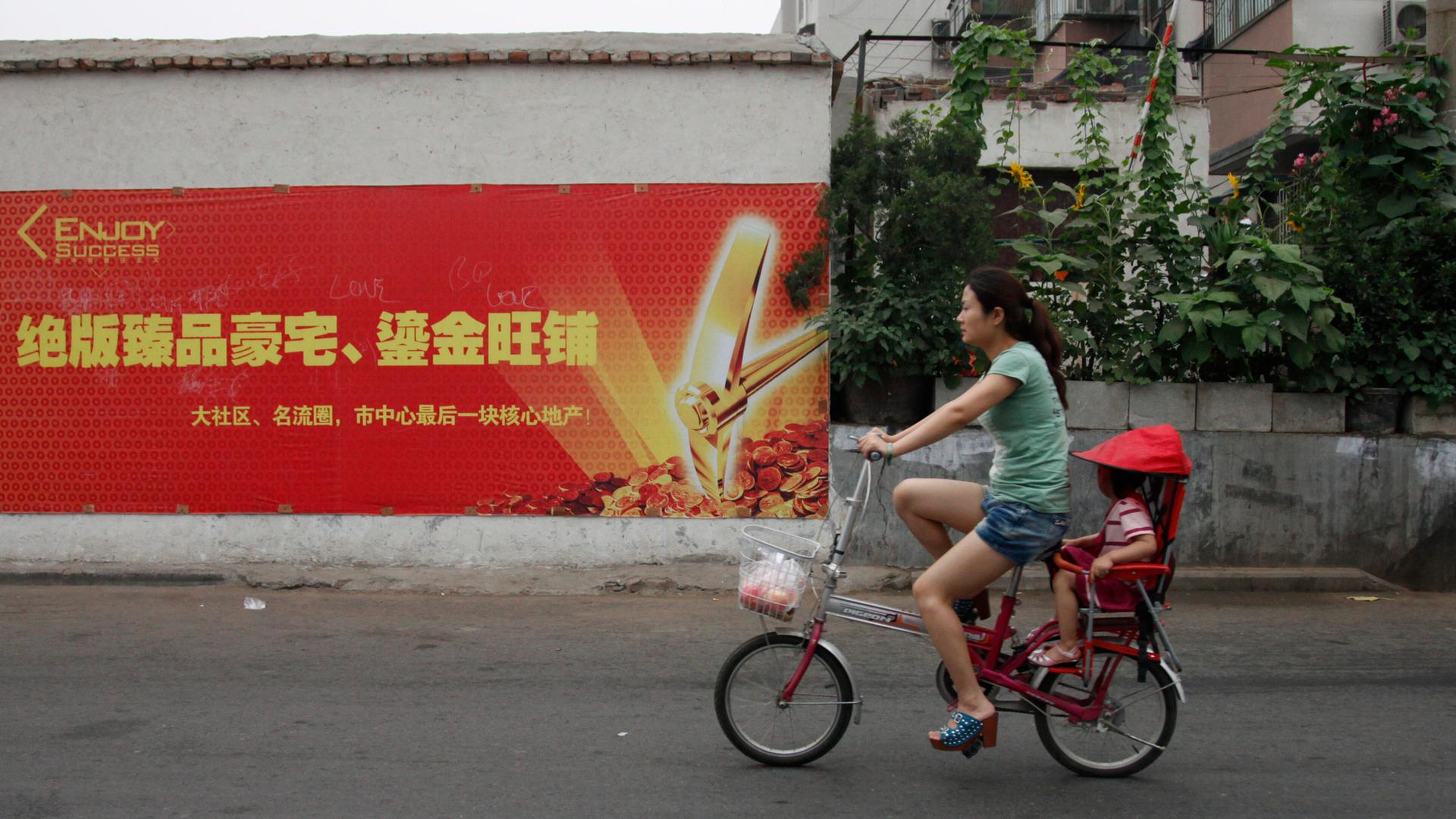 Eine Frau fährt in China auf einem kleinen Fahrrad eine Straße entlang, ein Kind sitzt hinten unter einem roten Sonnenschutz.
