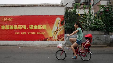 Eine Frau fährt in China auf einem kleinen Fahrrad eine Straße entlang, ein Kind sitzt hinten unter einem roten Sonnenschutz.
