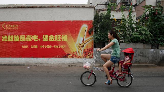 Eine Frau fährt in China auf einem kleinen Fahrrad eine Straße entlang, ein Kind sitzt hinten unter einem roten Sonnenschutz.
