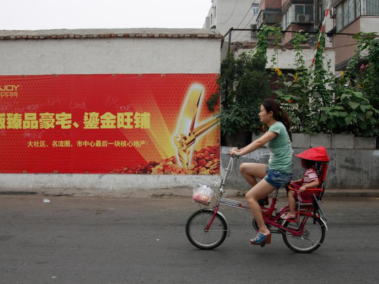 Eine Frau fährt in China auf einem kleinen Fahrrad eine Straße entlang, ein Kind sitzt hinten unter einem roten Sonnenschutz.
