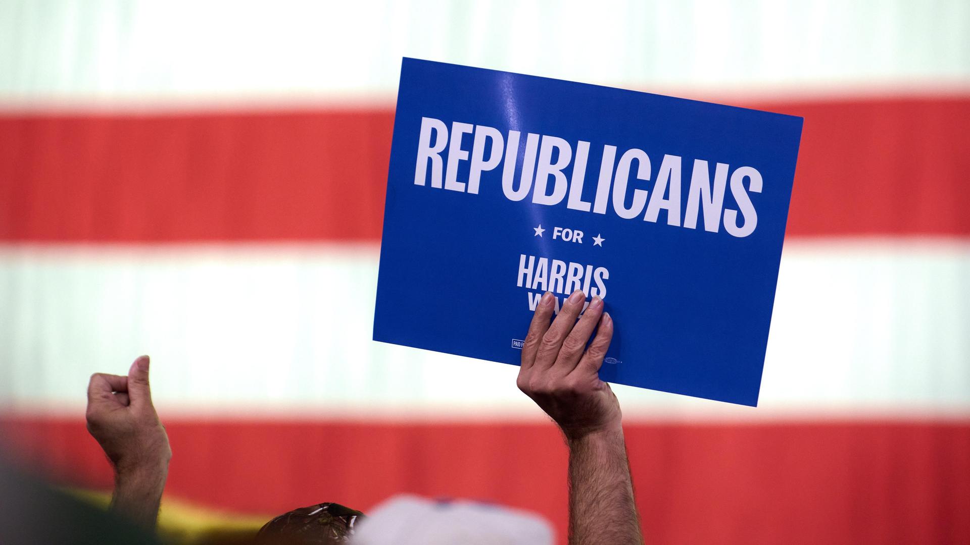 Vor der US-Flagge hält eine Person ein Schild hoch, auf dem "Republicans for Harris" zu lesen ist.