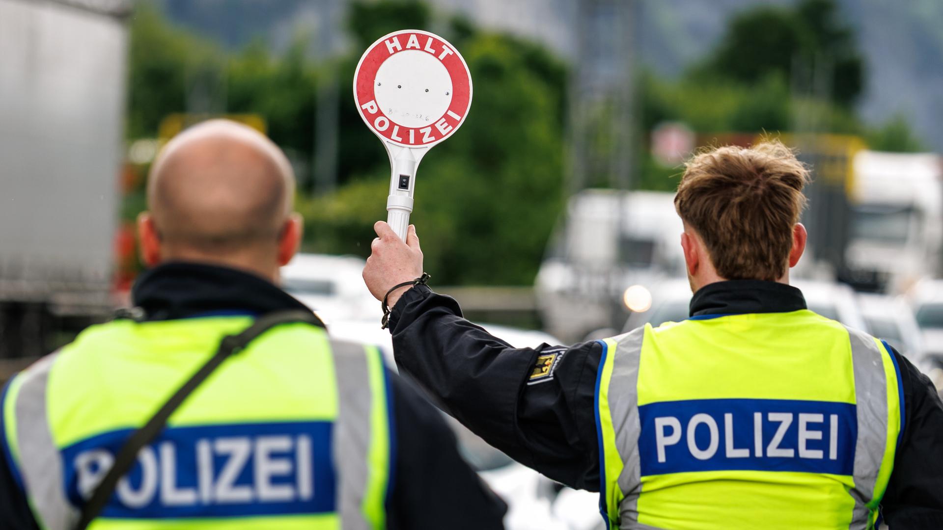 Zwei Polizisten stehen an einer Kontrollstelle an der deutschen Grenze. Einer von beiden hebt eine Kelle in die Höhe, auf der "Halt - Polizei" zu lesen ist.