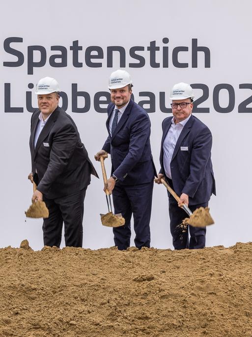 Spatenstich für das Rechenzentrum in Lübbenau. Männer im dunklen Anzug mit Helm auf dem Kopf und Schaufel in der Hand vor einem Schild "Spatenstich Lübbenau"  