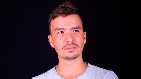Bob Blume steht im weißen T-Shirt vor einem dunklen Hintergrund. Unter dem Ärmel ist ein farbiges Tattoo auf seinem rechten Oberarm zu sehen.