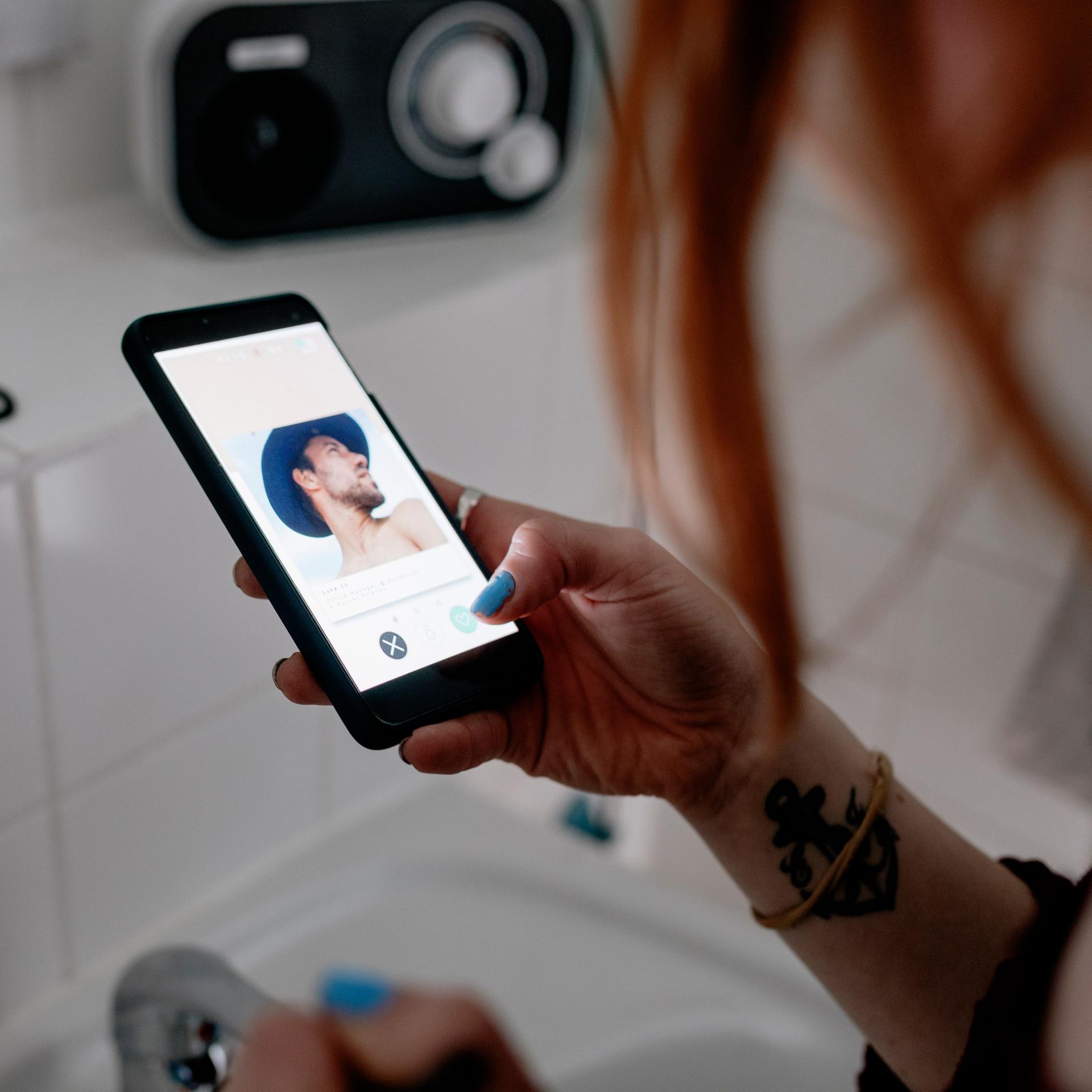 Eine Frau steht mit Schminkutensilien vor einem Badezimmerspiegel und benutzt eine Dating-App auf ihrem Smartphone.