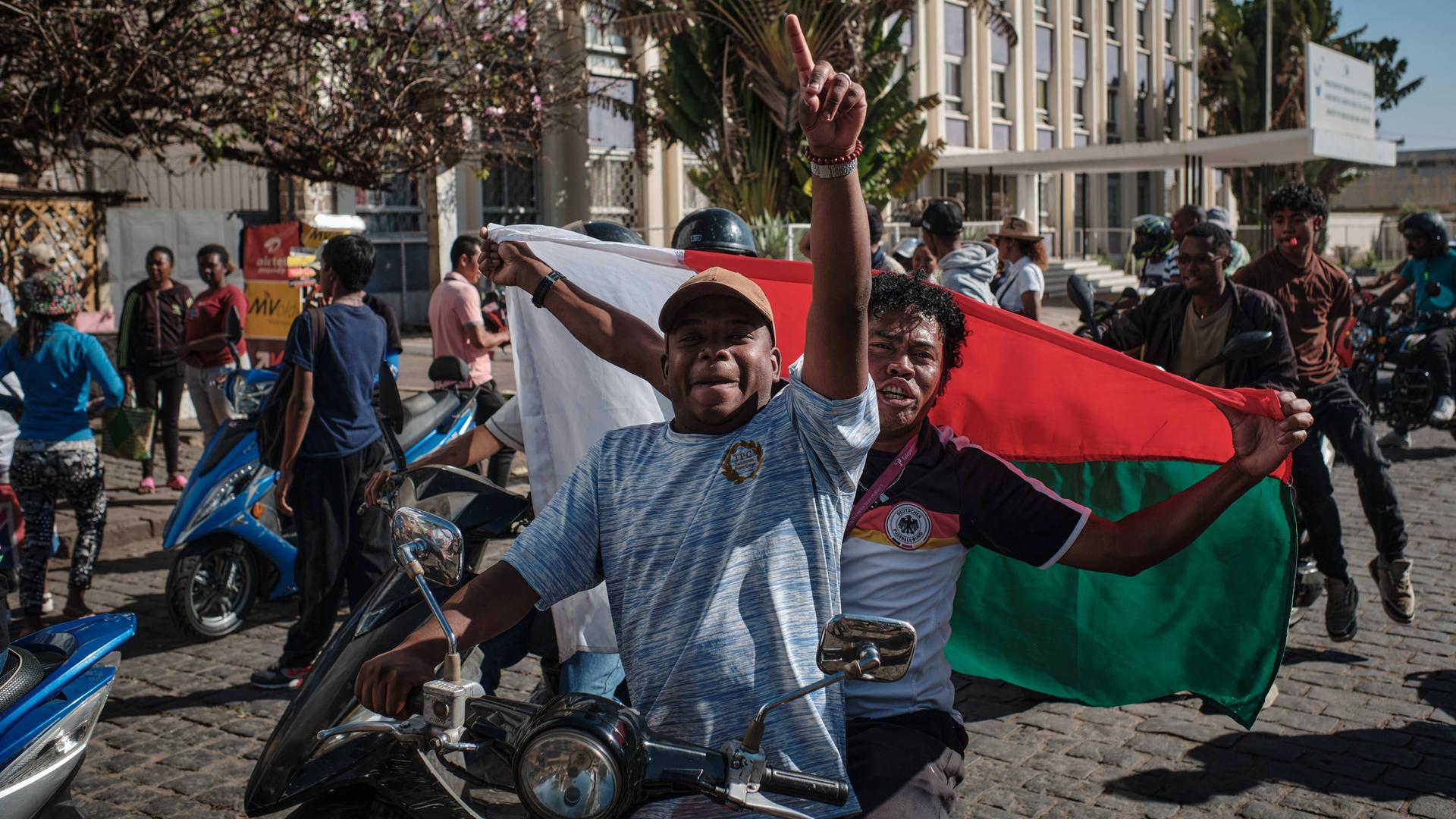 Junge Menschen auf kleinen Motorrädern und Motorrollern in Madagaskar. Sie schwenken Flaggen und protestieren gegen die Regierung.