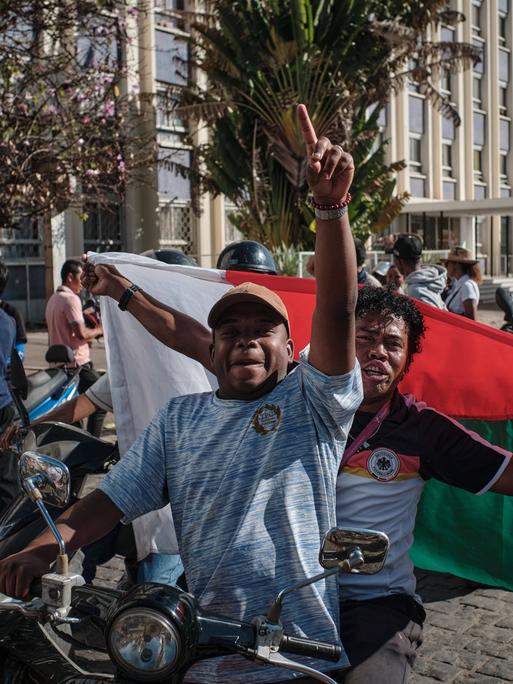 Junge Menschen auf kleinen Motorrädern und Motorrollern in Madagaskar. Sie schwenken Flaggen und protestieren gegen die Regierung.